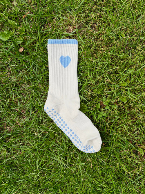 A single white ribbed sock with a light blue heart on the calf and blue grip dots on the sole, placed on green grass.