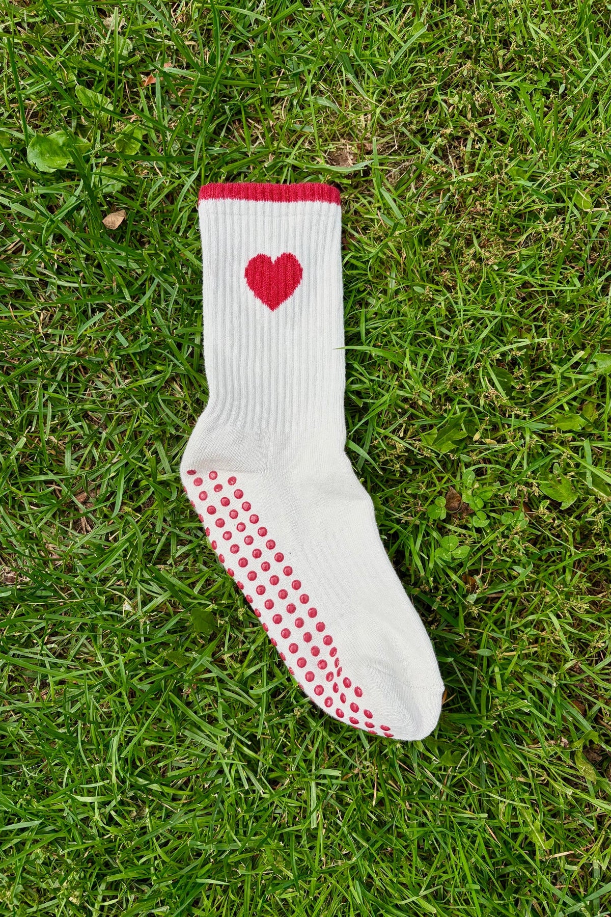A single white sock with a red heart on the cuff and red grip dots on the sole, laid flat on green grass.